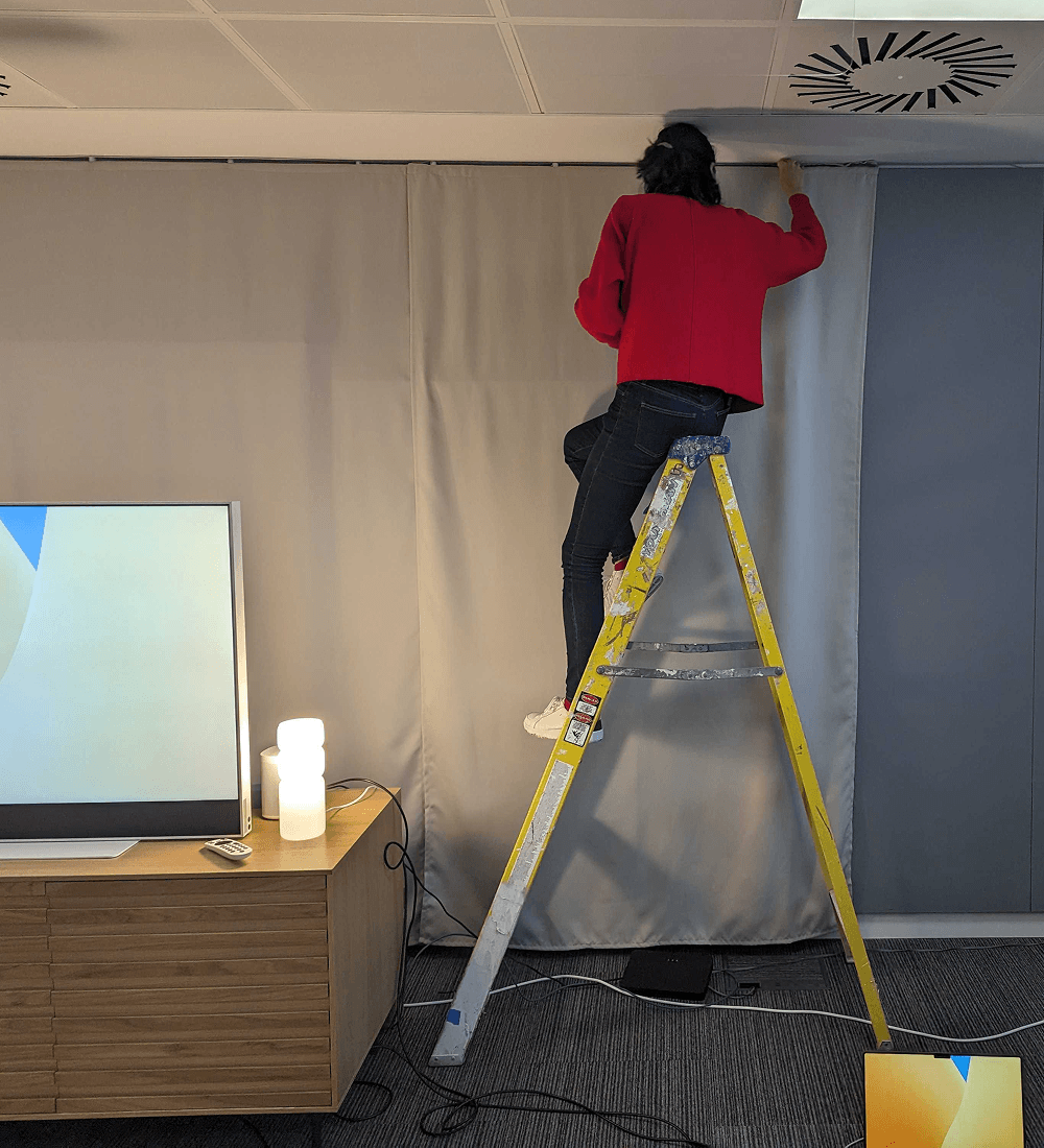 Lucrecia standing on a ladder, reaching up to finish hanging a curtain backdrop in an office space